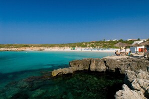 A coastal beach scene with clear turquoise waters, rocky formations, and a few beach umbrellas. - Villa Tama by Villa Plus (Sant Lluís)