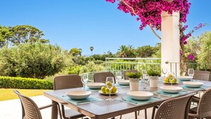 Uma área de refeições ao ar livre com uma mesa posta para quatro pessoas, rodeada por cadeiras, e uma pérgola com flores cor-de-rosa.