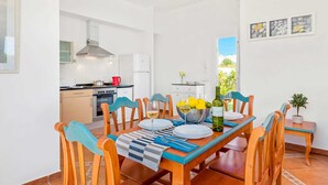 A modern kitchen with white cabinets, a stove, and a red kettle. There is a dining area with a table set for two, chairs, and a potted plant. - Villa Torrenit by Villa Plus (Alayor)