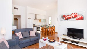 A modern living room with a grey sofa, a coffee table with fruits and magazines, a TV on a white stand, and a kitchen in the background. - Villa Torresoli by Villa Plus (Alayor)