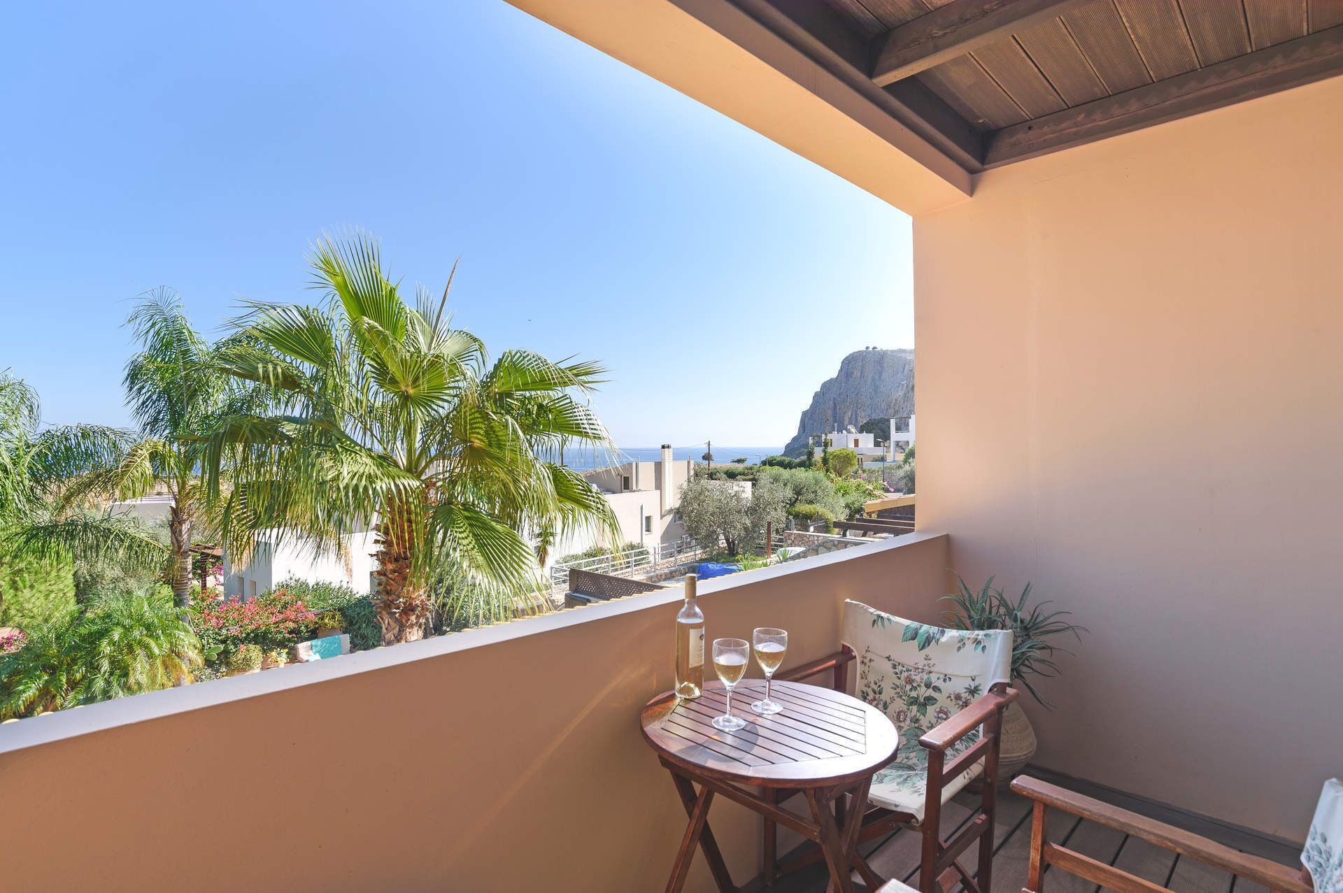 A balcony with a table and chairs, overlooking a coastal area with palm trees and a mountain in the distance.
