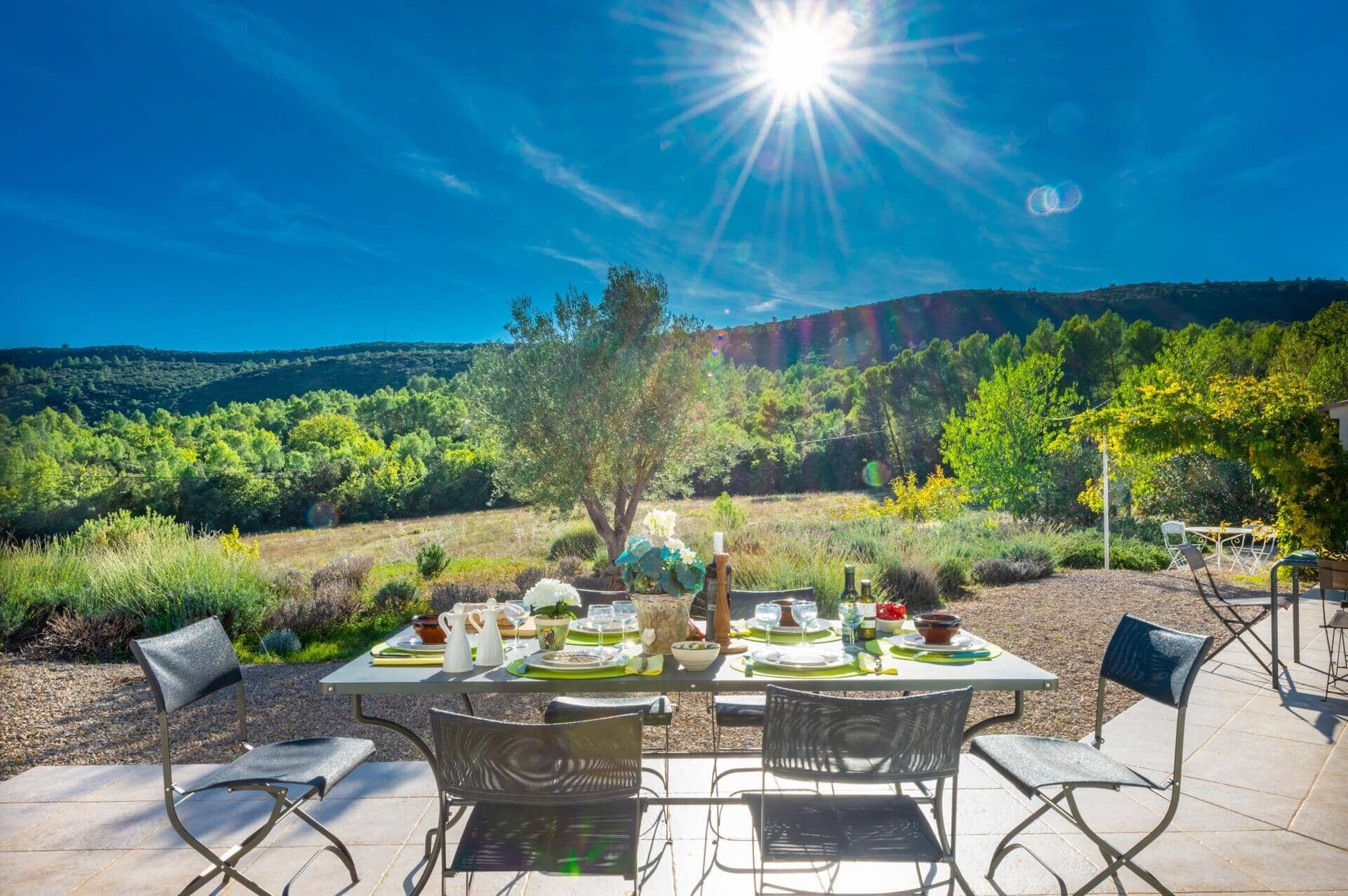 An outdoor dining area with a table set for four, surrounded by chairs, overlooking a scenic landscape with hills and trees.