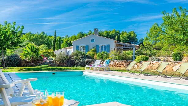 Ein Swimmingpool mit Liegestühlen und einem Tisch mit Getränken, umgeben von Grünpflanzen und einem Haus im Hintergrund.