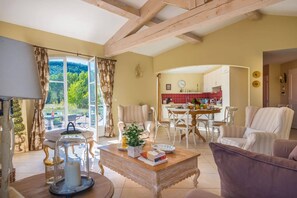 A cozy living room with a dining area, a kitchen in the background, and a large window offering a view of greenery.
