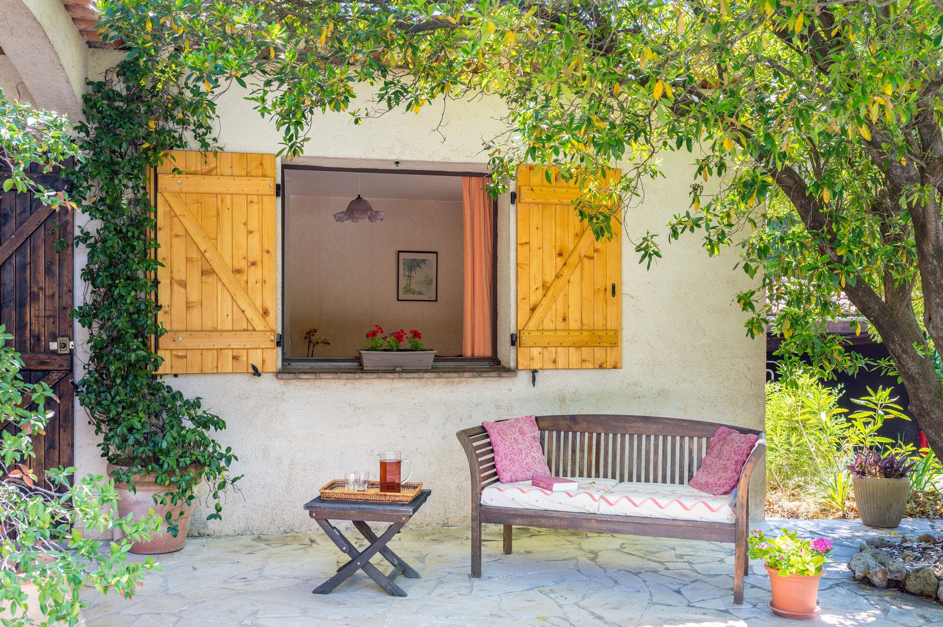 A cozy outdoor seating area with a bench and a small table, surrounded by potted plants and a rustic building with wooden shutters.