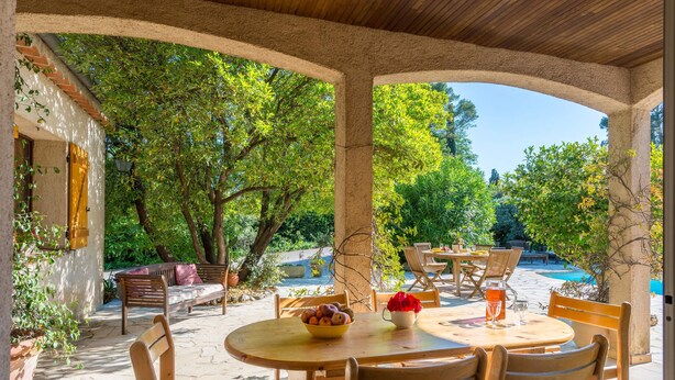 Eine Terrasse im Freien mit einem runden Holztisch, vier Holzstühlen, einer Obstschale und einer Vase mit roten Blumen.
