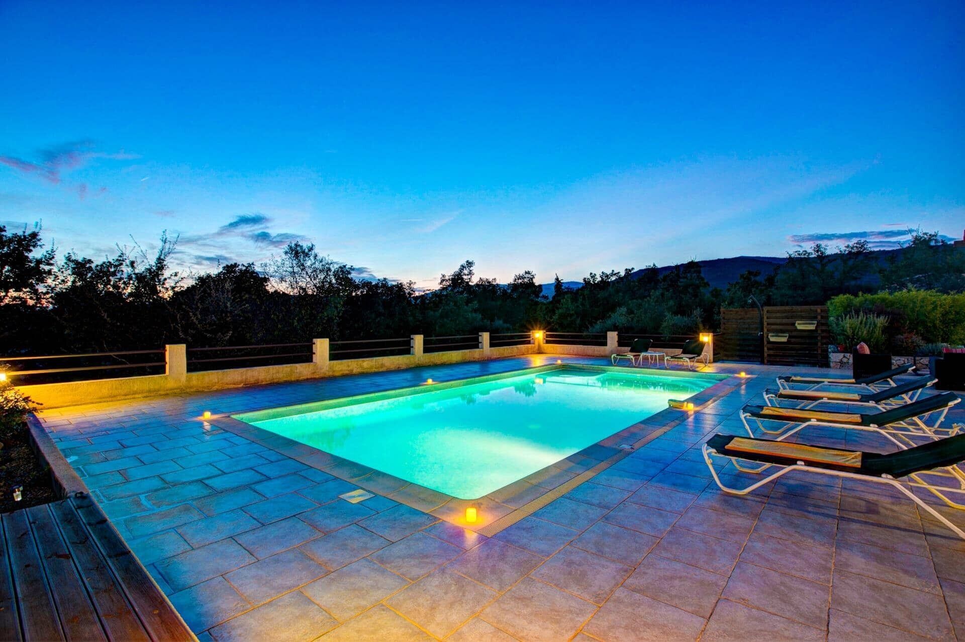 A rectangular swimming pool with a tiled deck, surrounded by a wooden fence and a wooden deck with lounge chairs.