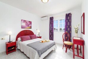 Un dormitorio con una cama roja, una mesita de noche con una lámpara, una silla, un escritorio con un espejo y una ventana con cortinas moradas.