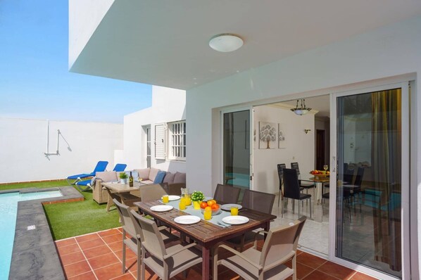 Un área de comedor al aire libre con una mesa dispuesta para cuatro personas, rodeada de sillas, cerca de una piscina y un porche cubierto.