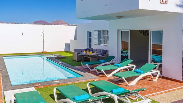 Un espace piscine doté de chaises longues et d’une table, entouré d’un bâtiment blanc offrant une vue sur les montagnes.