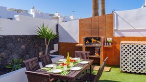 Un espace extérieur aménagé pour les repas, avec un barbecue, un mur en pierre et des palmiers.