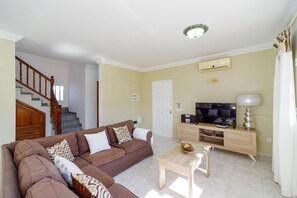 Una sala de estar con un sofá marrón, una mesa de centro de madera, un televisor sobre un mueble de madera y una escalera que conduce a otra habitación.