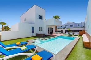 Uma área moderna à beira da piscina, com piscina retangular, espreguiçadeiras e um edifício branco com telhado plano.