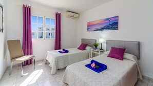 Ein Hotelzimmer mit zwei Betten, einem Sessel, einem Fenster mit Vorhängen, einer Klimaanlage, einem Nachttisch mit Lampe, einem Gemälde und einer Topfpflanze.