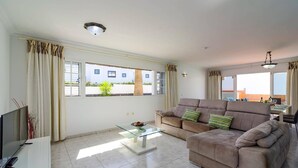 Ein Wohnzimmer mit einer Couch, einem Glastisch und einem Fernseher. Es gibt Fenster mit Vorhängen, eine Deckenleuchte und einen Blick auf ein Gebäude.