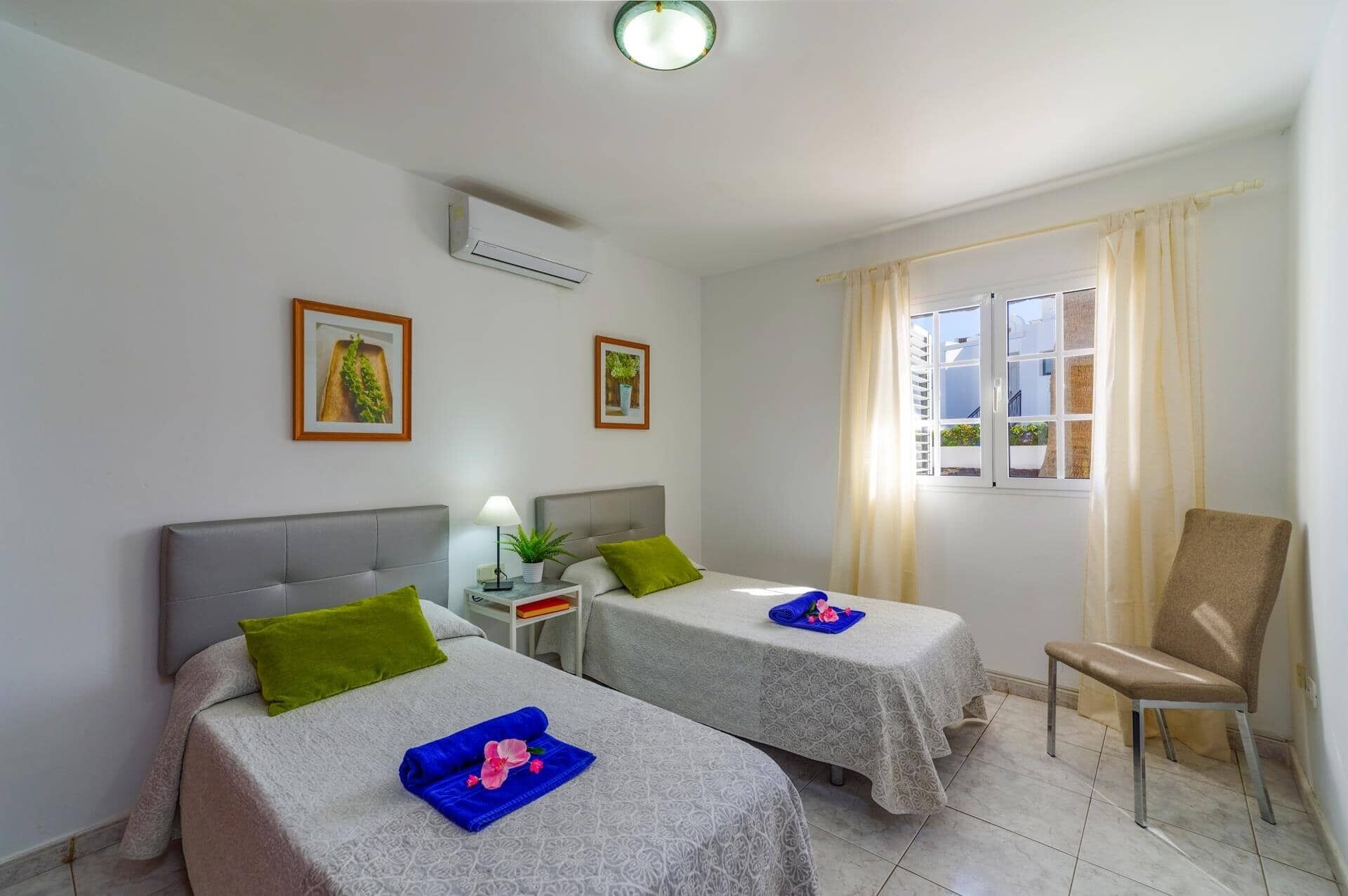 A hotel room with two beds, a chair, a window with curtains, and a ceiling fan.