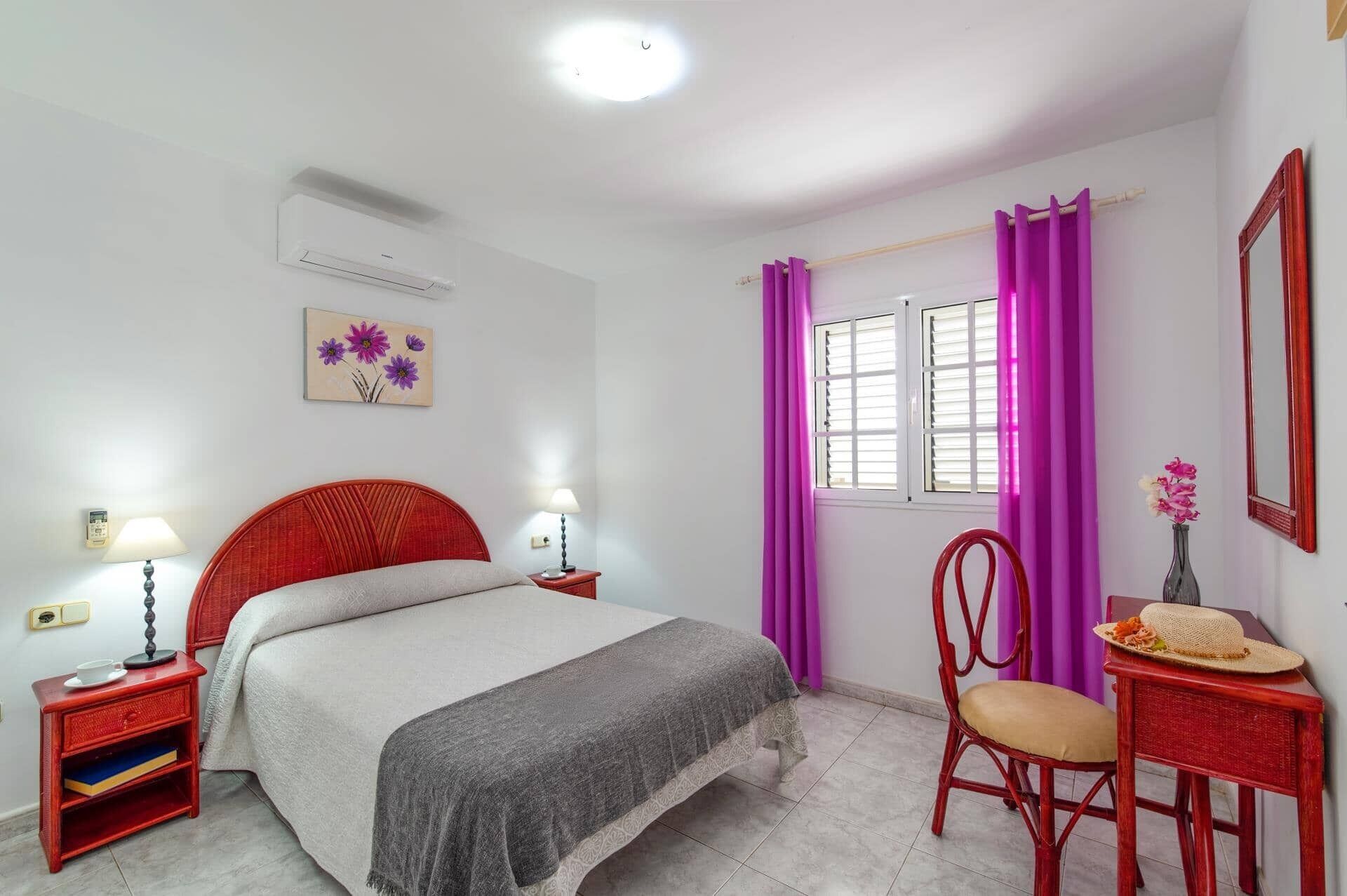 Un dormitorio con una cama, dos mesitas de noche con lámparas, una silla, un escritorio con un florero, una ventana con cortinas moradas y un espejo instalado en la pared.