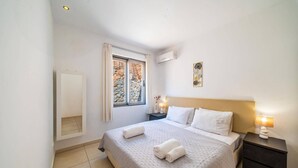 Ein ordentlich eingerichtetes Schlafzimmer mit einem Bett, Nachttischen, einem Fenster mit Vorhängen und einer Wand-Klimaanlage.