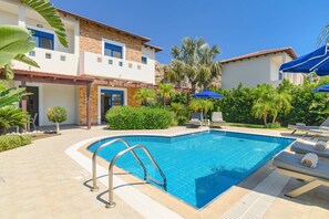 Ein Swimmingpool mit Metallleiter, umgeben von einer Terrasse mit Liegestühlen und Sonnenschirmen.