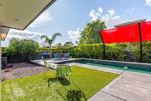 A backyard with a swimming pool, a patio with outdoor furniture, and a red sunshade.
