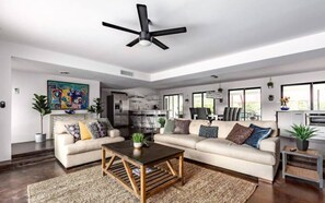 A spacious living room with a large ceiling fan, a sofa set, a coffee table, and a kitchen area visible through an open doorway. - Designer 5 Bd Home/Fountain Pool/ Spa/ Game Patio (Phoenix)
