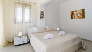 A bedroom with a bed, two bedside tables, a window with sheer curtains, and two wall-mounted paintings. - Villa Despina by Villa Plus (Pefki)