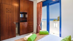 Quarto de hotel com guarda-roupa de madeira, cama com toalhas verdes e bege, e varanda com guarda-corpo azul com vista para o mar.