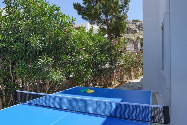 A blue ping pong table with a net in a garden area, surrounded by various green plants and trees.
