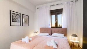 A neatly made bed with two pillows and two rolled towels, a window with curtains, and two framed artworks on the wall. - Villa Maya by Villa Plus (Pefki)