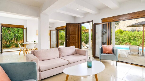 A spacious living area with a sofa, two armchairs, and a coffee table. Large glass doors open to a patio with a pool and greenery. - Villa Maya by Villa Plus (Pefki)