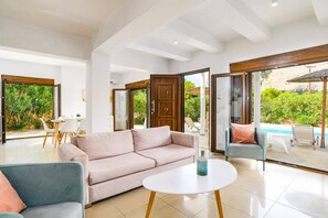 A spacious living area with a sofa, two armchairs, and a coffee table. Large glass doors open to a patio with a pool and greenery.
