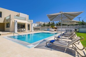 A swimming pool with lounge chairs and umbrellas, a patio area with a table and chairs, and a modern building with a glass door.