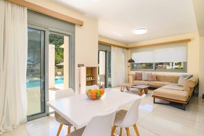 A modern living room with a sofa, a dining table with chairs, and a view of a swimming pool through glass doors.