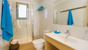 Et moderne badeværelse med brusekabine i glas, et hvidt toilet, en vask med spejl og blå håndklæder.