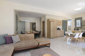 A modern living room with a sofa, a dining area with a table and chairs, and a kitchen with cabinets and a refrigerator. - Villa Paleos Evergreen by Villa Plus (Ialysos)
