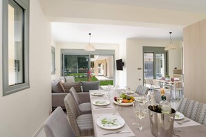 A dining area with a table set for four, a sofa, and a view of a garden. - Villa Paleos Sunset by Villa Plus (Ialysos)