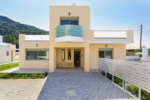 A modern two-story house with a balcony, a paved entrance, and a view of a green hillside. - Villa Paleos Sunset by Villa Plus (Ialysos)