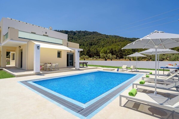 A modern pool area with a rectangular pool, lounge chairs, and umbrellas. - Villa Paleos Sunset by Villa Plus (Ialysos)