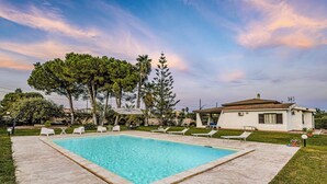 A swimming pool with lounge chairs, a white building, and trees in the background. - Villa Rosanna by Villa Plus (Fontane Bianche)
