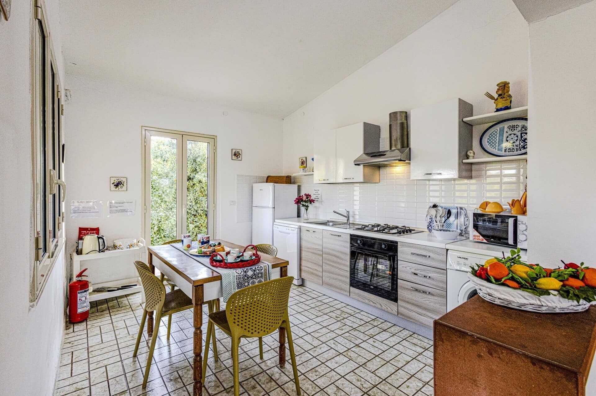 Une cuisine compacte avec des meubles blancs, une cuisinière à gaz, un four à micro-ondes et un évier. On y trouve une table à manger avec des chaises et une fenêtre donnant sur un paysage verdoyant.