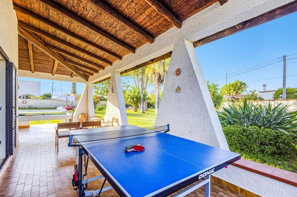 Una mesa de ping pong al aire libre con red, rodeada por un porche cubierto con vigas de madera y vista a un jardín.