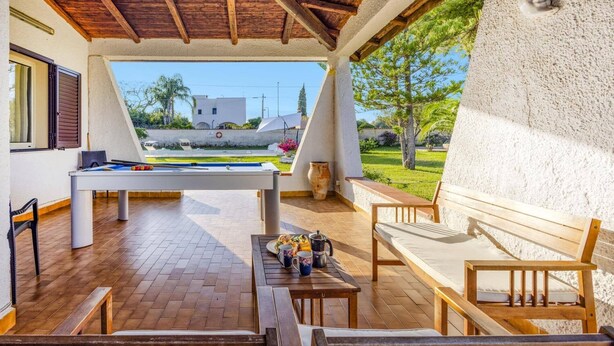 A covered outdoor area with a pool table, seating, and a view of a pool and greenery. - Villa Rosanna by Villa Plus (Fontane Bianche)