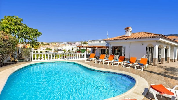 A swimming pool with lounge chairs, a white building with a tiled roof, and a clear blue sky.