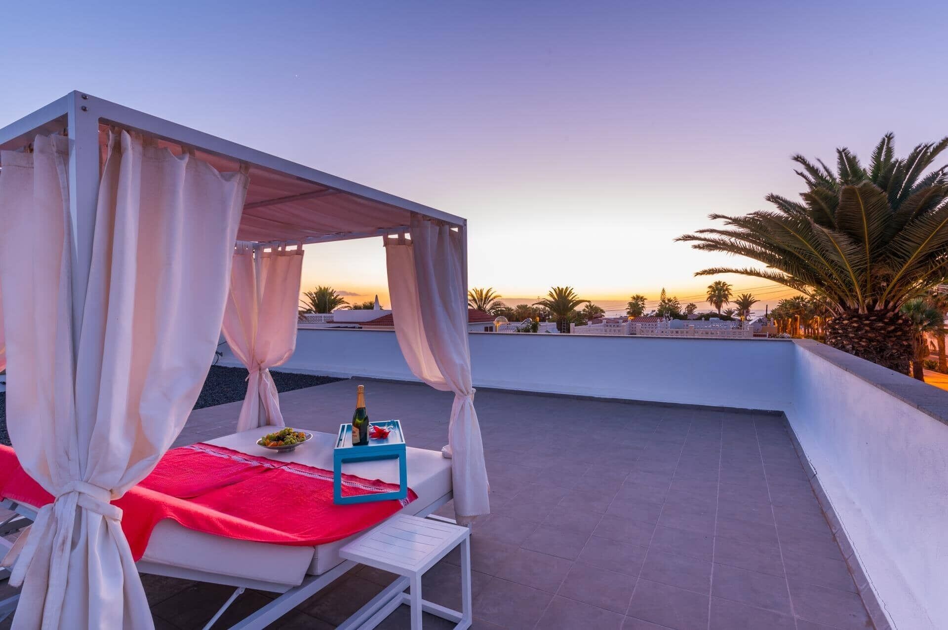 A rooftop terrace with a covered seating area, a table with a bottle and a bowl of fruit, and a view of palm trees and buildings at sunset.