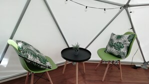 A room with a wooden floor, two green chairs with leaf-patterned cushions, a small black table with a potted plant, and string lights on a white wall. - Escapada a Tortuguero  (Guapiles)