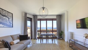 Un salon spacieux comprenant un canapé, une table à manger et des chaises, une grande fenêtre offrant une vue sur les montagnes et un luminaire suspendu.