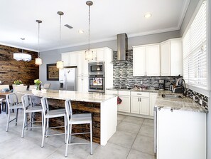 A modern kitchen with white cabinets, a large island with bar stools, stainless steel appliances, and a stone backsplash.