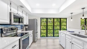 A modern kitchen with white cabinetry, stainless steel appliances, and a large window providing a view of greenery. - Sunset Captiva 10 (Captiva)