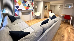 A modern living room with a sectional sofa, a colorful geometric wall art, a wooden floor, and a desk with a chair. - Stylish Studio Apartment in Clifton Village (Bristol)