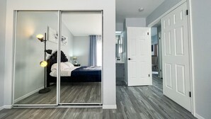 Slaapkamer met een schuifdeurkast, een bed met wit beddengoed, een nachtkastje met een lamp en uitzicht op een andere kamer.